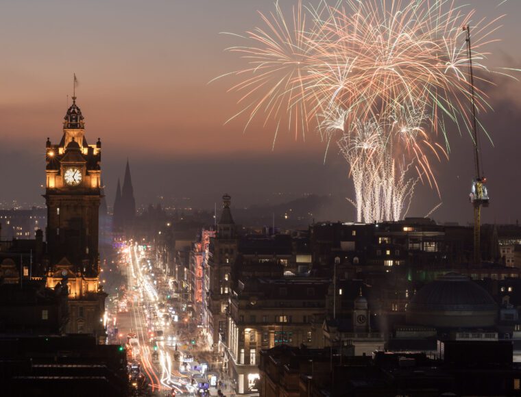 View of Princess Street, Edinburgh, from Calton Hill at dusk, with lights on and firework display in the sky.